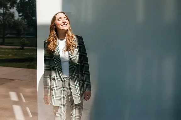 Eine Frau mit langen Haaren, die einen quadratischen Blazer und eine Hose trägt, steht an einer weißen Wand und blickt mit einem freudigen Ausdruck nach oben.