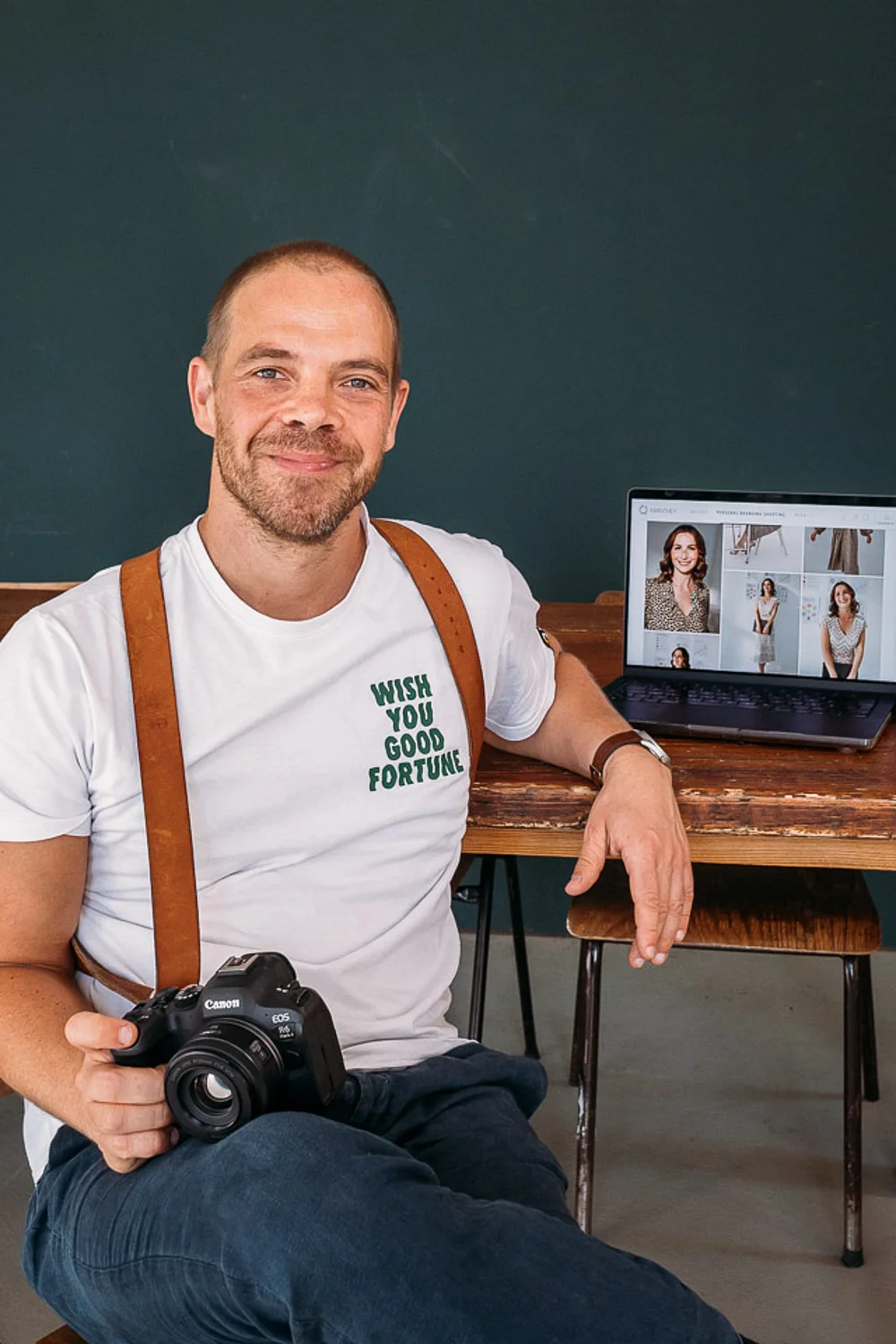 Christian Manthey im weißen T-Shirt mit Kamera vor einer dunkelgrünen Wand, daneben ein Laptop mit Porträts