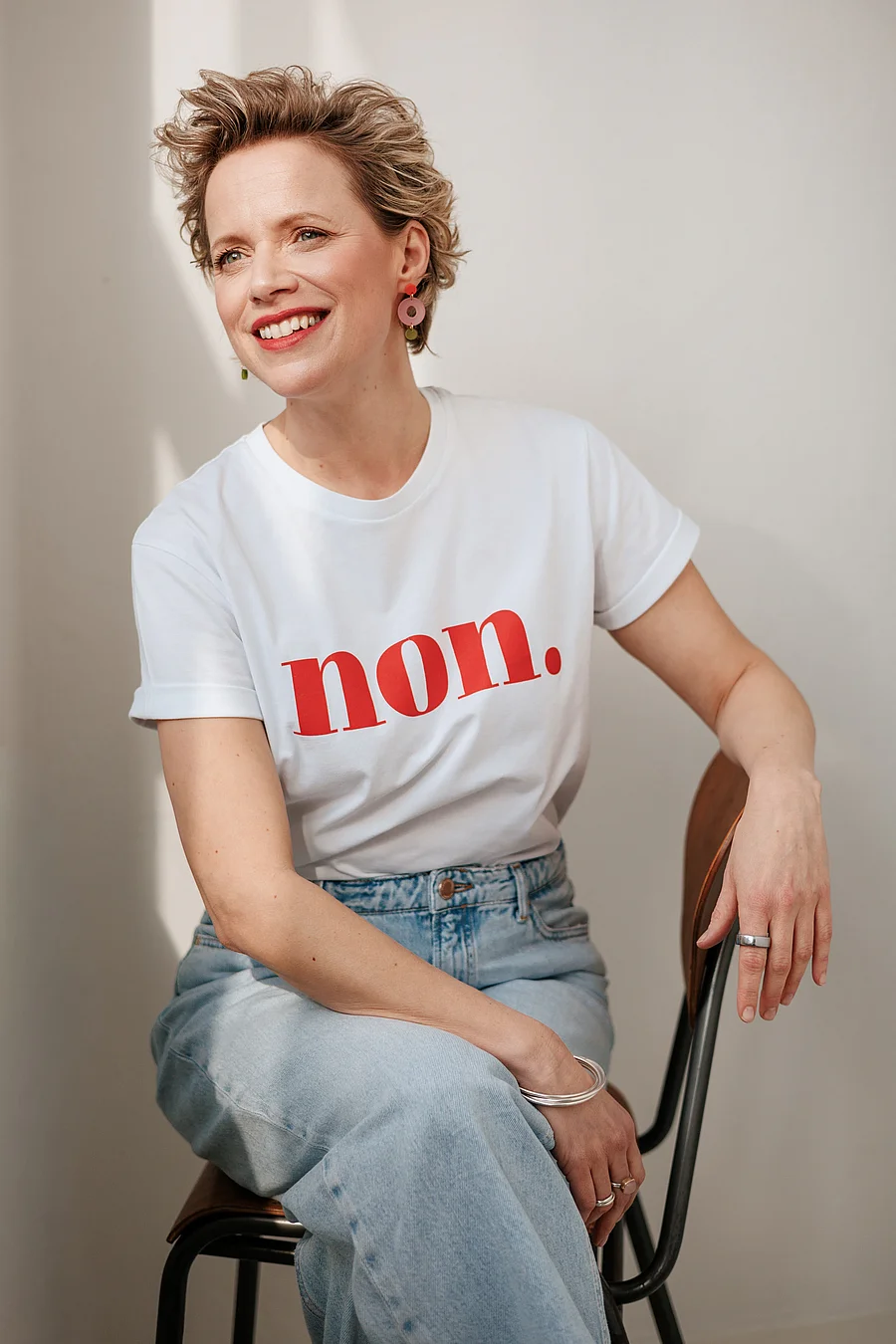 Frau mit kurzen Haaren im weißen Shirt mit rotem Schriftzug sitzt auf einem Stuhl und lacht in die Kamera