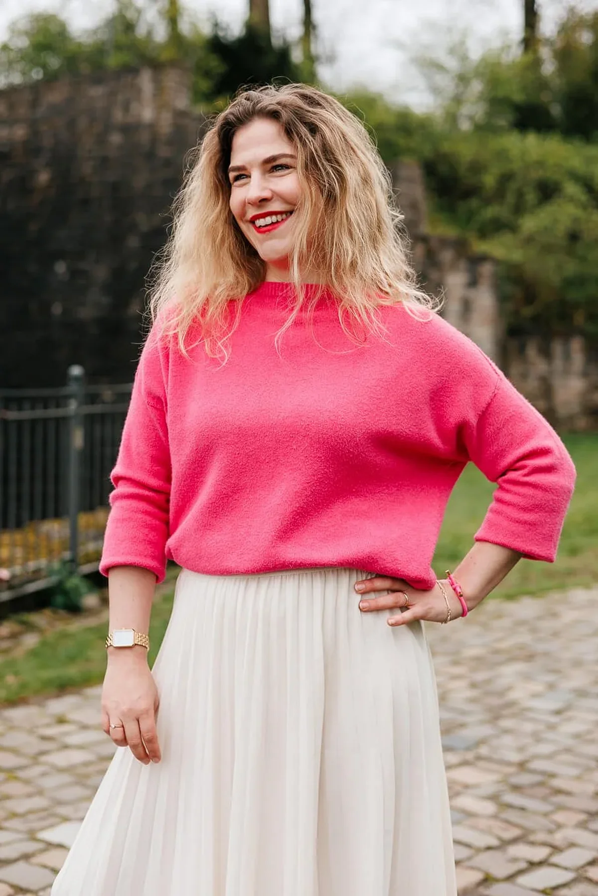 Frau mit blonden Locken im pinken Pullover und weißem Rock lacht, eine Hand in der Hüfte, auf einem Kopfsteinpflasterweg