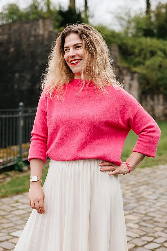 Eine Frau mit lockigem blondem Haar trägt einen rosa Pullover und einen weißen Rock und steht draußen vor einer Steinmauer mit Grün im Hintergrund.