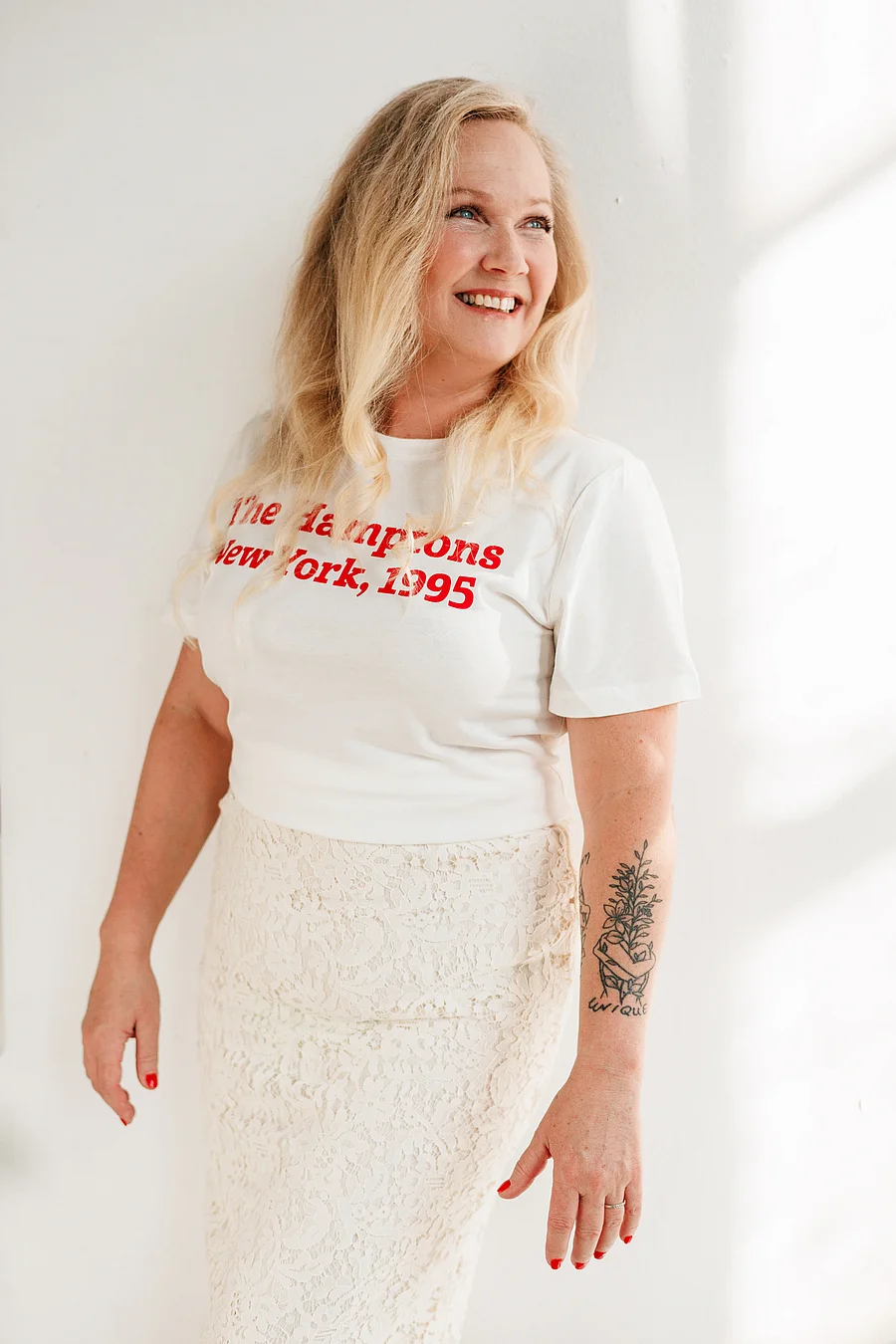 Blonde Frau im weißen Band-T-Shirt und weißem Rock steht lachend vor einer hellen Wand, Tattoo am Unterarm