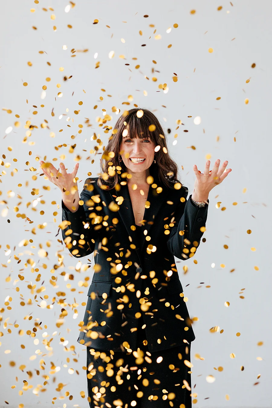 Frau im schwarz-goldenen Kleid wirft goldenes Konfetti in die Luft und lacht, helles Studio