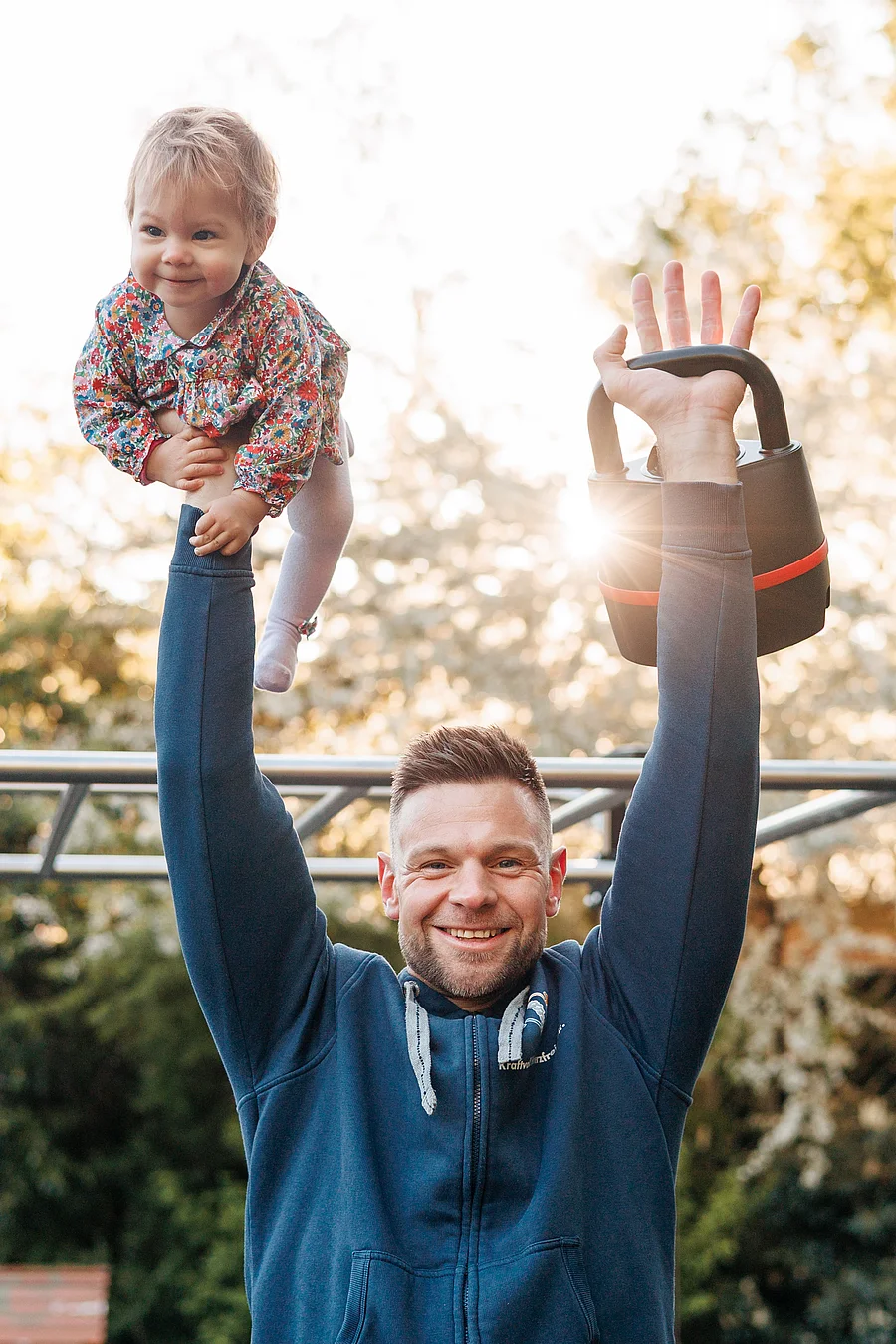 Mann in blauer Jacke stemmt draußen eine Kettlebell und hält ein Kleinkind in die Luft, beide lachen