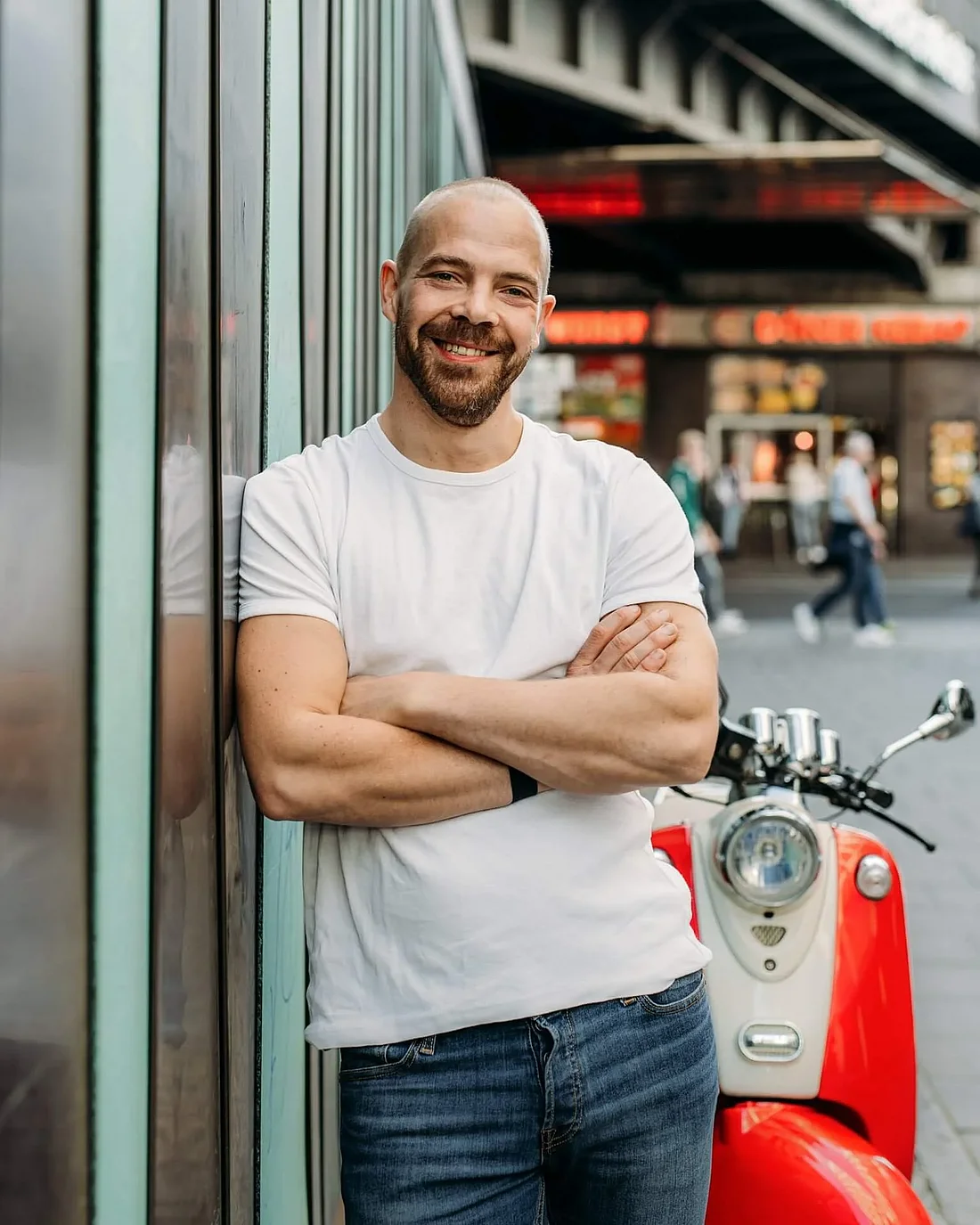 Christian Manthey im weißen T-Shirt lehnt lachend an einer Hauswand in Berlin, daneben ein roter Motorroller