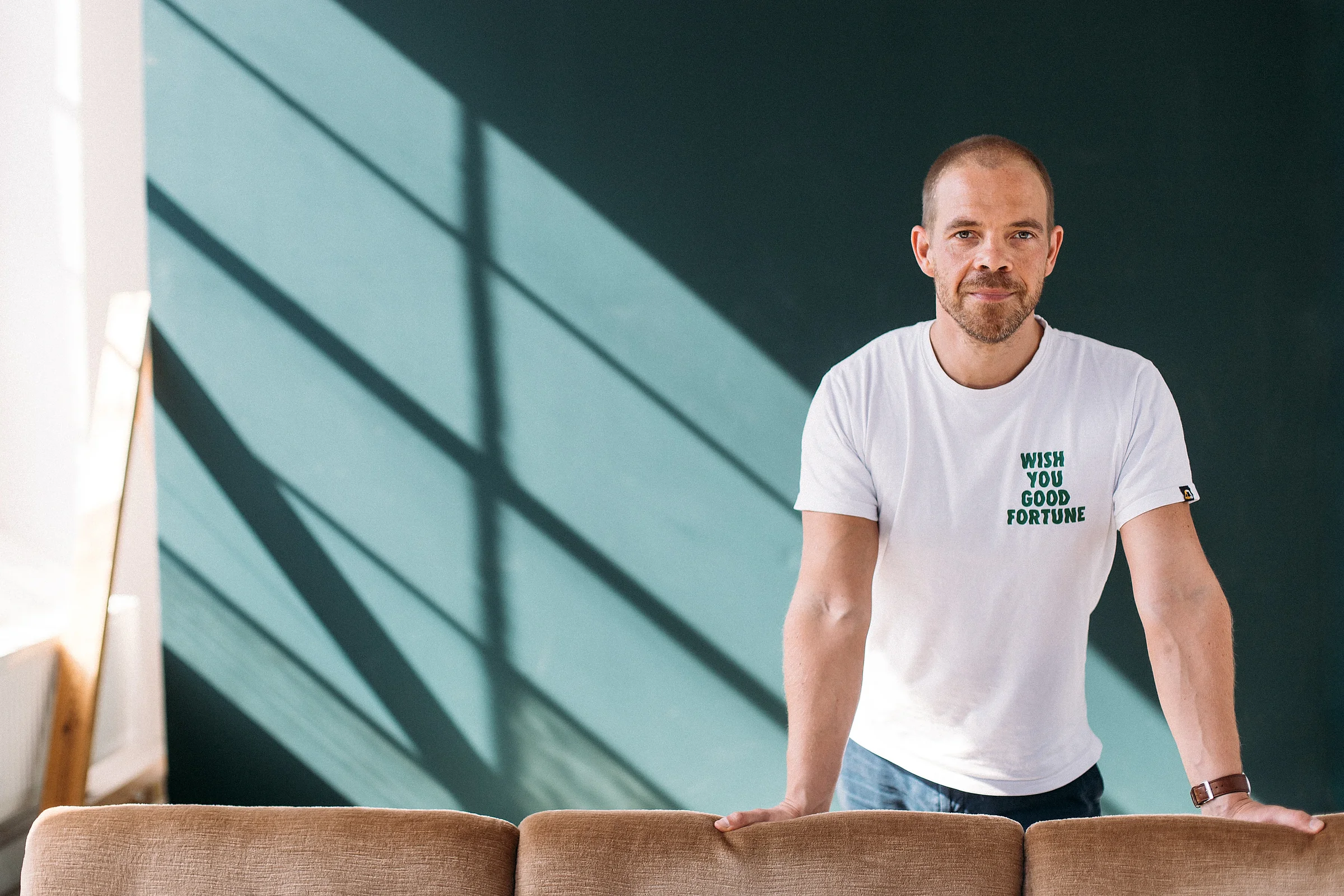 Christian Manthey im weißen T-Shirt stützt sich auf ein Sofa, dahinter eine dunkelgrüne Wand mit Fensterschatten