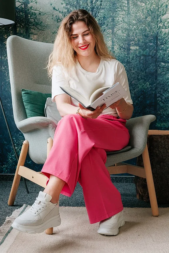 Eine Frau mit lockigen Haaren, die ein weißes T-Shirt und eine rosa Hose trägt, sitzt auf einem Stuhl und liest ein Buch mit einem Waldwandbild im Hintergrund.