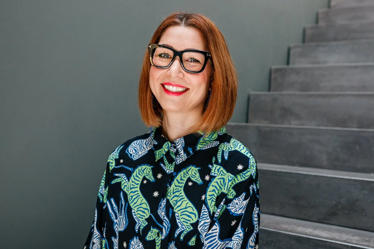 Frau mit roten Haaren, schwarzer Brille und gemusterter Bluse lacht vor einer grauen Treppe